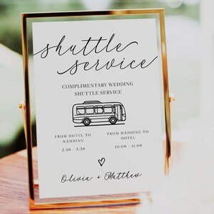 May include: A gold-framed sign with white background and black text announcing a complimentary wedding shuttle service. The sign includes a simple illustration of a bus and the shuttle times: 2:30-3:30 from hotel to wedding and 10:00-11:00 from wedding to hotel. The sign is signed "Olivia + Matthew".