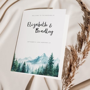 Op de afbeelding: Een wit huwelijksboekje met een aquarel-illustratie van een bergketen en dennenbomen. De tekst luidt "Welkom op de bruiloft van Elizabeth & Bradley, 21 september 20XX | Monterey, CA."
