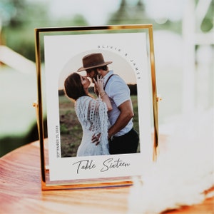 Puede incluir: Una tarjeta de número de mesa blanca con un marco dorado. La tarjeta tiene una foto de una pareja besándose bajo un arco. El texto en la tarjeta dice "Olivia & James, 25 de septiembre de 20XX, Mesa Dieciséis."