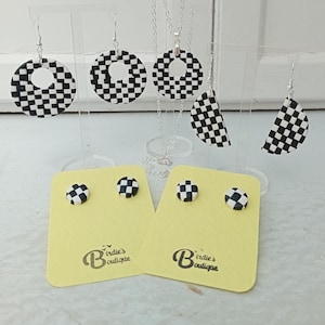 May include: Checkerboard pattern jewelry set with a necklace, earrings, and stud earrings. The necklace and larger earrings are round with a hole in the center. The smaller earrings are round and solid. The jewelry is displayed on a white stand with yellow cards that say "Birdie's Boutique".