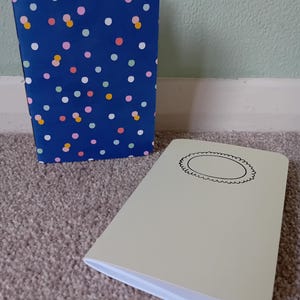 May include: Two small notebooks. One has a blue cover with a colorful polka dot pattern. The other has a light beige cover with a black circle with a decorative border.