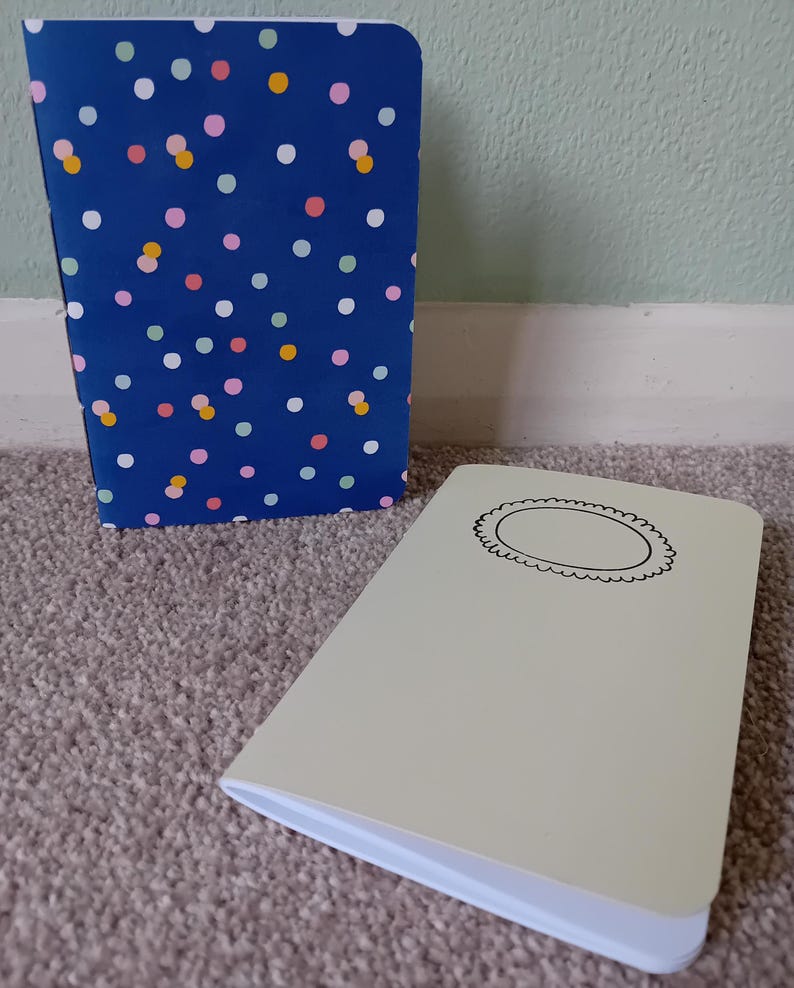 May include: Two small notebooks. One has a blue cover with a colorful polka dot pattern. The other has a light beige cover with a black circle with a decorative border.