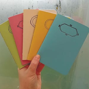 May include: Five small notebooks with different colored covers. The covers are green, pink, cream, yellow, and blue. Each notebook has a simple design on the front.
