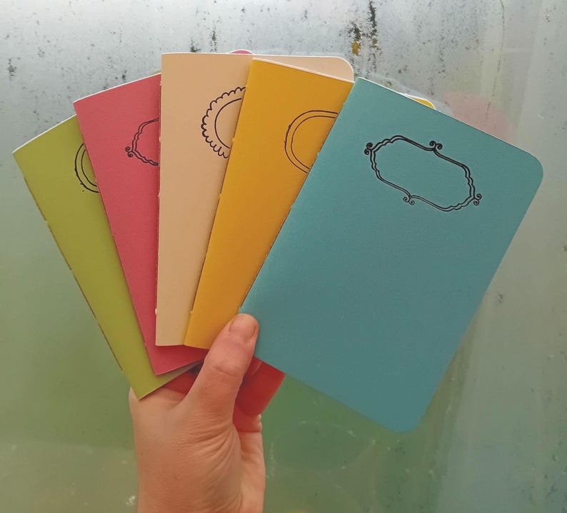 May include: Five small notebooks with different colored covers. The covers are green, pink, cream, yellow, and blue. Each notebook has a simple design on the front.