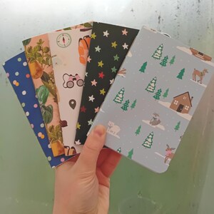 May include: A stack of five small, colorful notebooks with different patterns. The notebooks have a variety of designs, including stars, trees, and a compass. The notebooks are perfect for journaling, note-taking, or sketching.