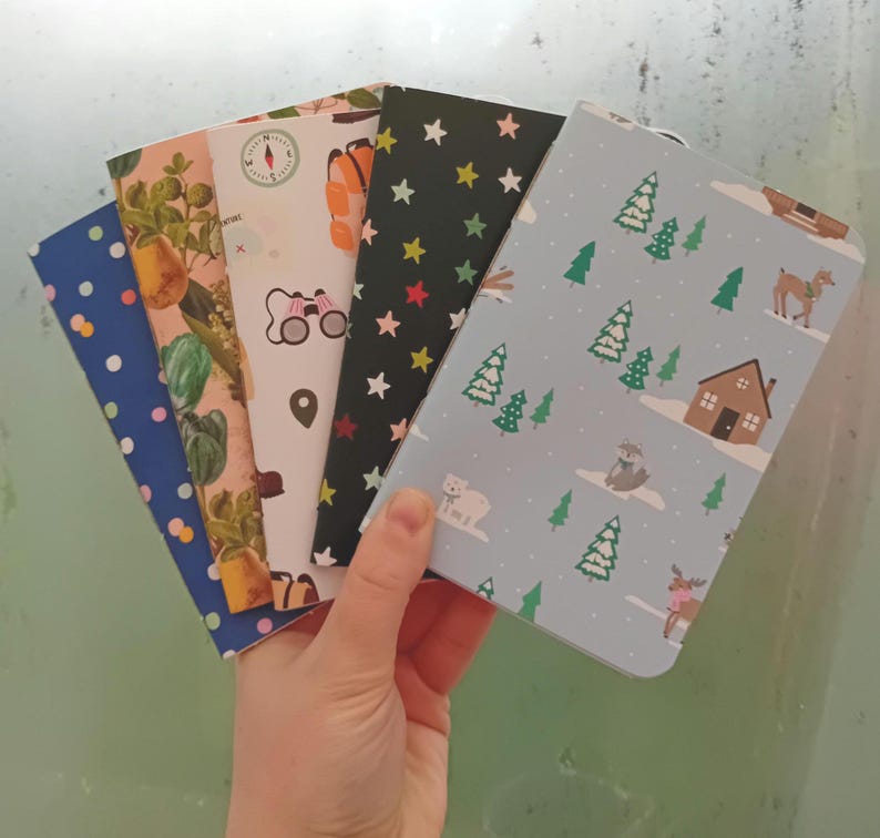 May include: A stack of five small, colorful notebooks with different patterns. The notebooks have a variety of designs, including stars, trees, and a compass. The notebooks are perfect for journaling, note-taking, or sketching.