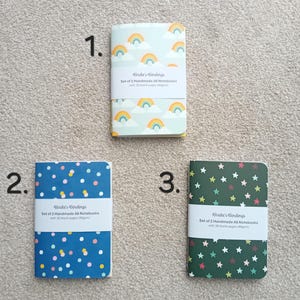 May include: Three sets of A6 notebooks with different designs. The first set has a rainbow and cloud design on a light blue background. The second set has a blue background with white and pink polka dots. The third set has a green background with yellow stars. Each set includes the text "Birdie's Bindings" and "Set of 2 Handmade A6 Notebooks with 30 blank pages (90gsm)".