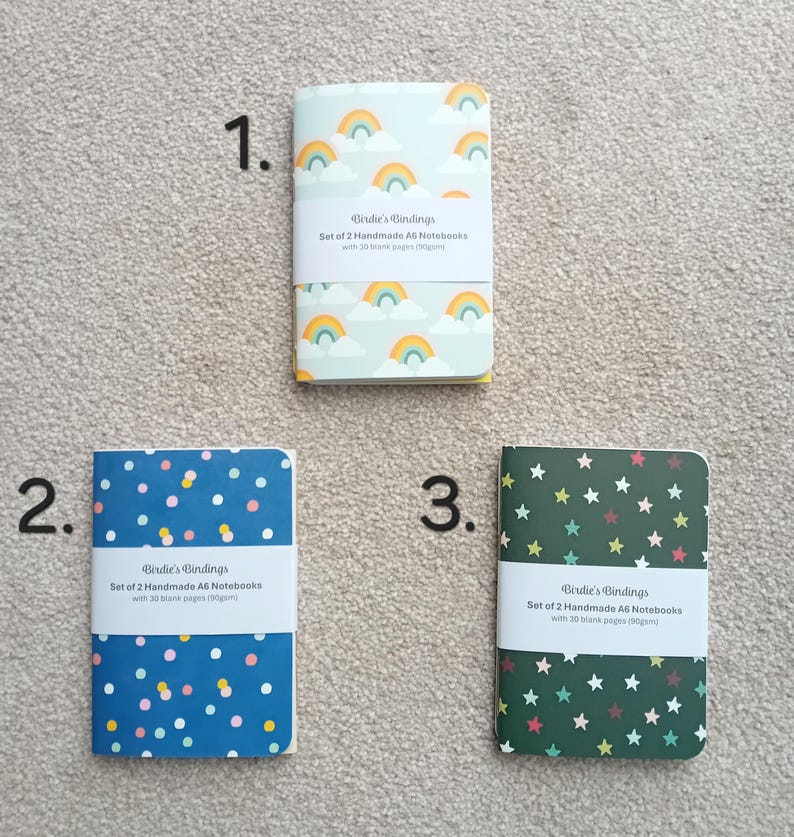 May include: Three sets of A6 notebooks with different designs. The first set has a rainbow and cloud design on a light blue background. The second set has a blue background with white and pink polka dots. The third set has a green background with yellow stars. Each set includes the text "Birdie's Bindings" and "Set of 2 Handmade A6 Notebooks with 30 blank pages (90gsm)".