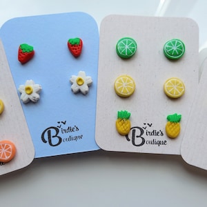 May include: Four sets of colourful fruit-themed stud earrings on display cards. Each set includes three pairs of earrings featuring a different fruit design: lime, lemon, pineapple, strawberry, and a white flower.