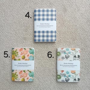 May include: Set of two A6 notebooks with floral designs. The notebooks have 30 blank pages and are 80gsm. The covers feature a floral pattern with pink, blue, and green colors. The text on the cover reads "Birdie's Bindings Set of 2 Handmade A6 Notebooks with 30 blank pages (80gsm)".