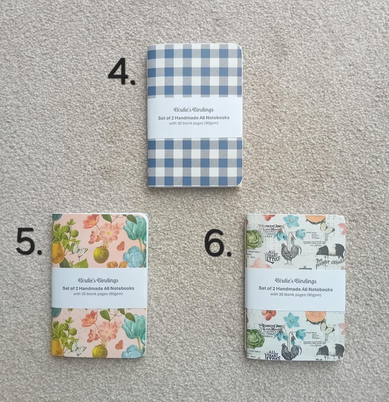 May include: Set of two A6 notebooks with floral designs. The notebooks have 30 blank pages and are 80gsm. The covers feature a floral pattern with pink, blue, and green colors. The text on the cover reads "Birdie's Bindings Set of 2 Handmade A6 Notebooks with 30 blank pages (80gsm)".