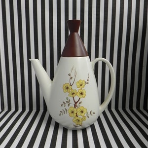 May include: A white ceramic teapot with a brown lid and a floral design featuring yellow flowers and green leaves. The teapot has a spout and a handle.