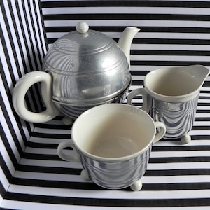 Może przedstawiać: Zestaw do herbaty w kolorze srebrnym i białym, składający się z czajnika, dzbanka na śmietanę i cukiernicy. Czajnik ma okrągły korpus i pokrywkę z gałką. Dzbanek na śmietanę i cukiernica mają pasiasty wzór.