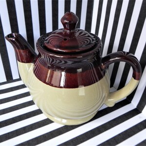 May include: A brown and beige ceramic teapot with a lid and a handle. The teapot is sitting on a black and white striped surface.