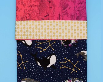 May include: A folded fabric item with three distinct patterned sections. The top section features pink clouds and birds, the middle has a gold floral design, and the bottom displays orcas and constellations on a navy background. The item is on a light blue surface.