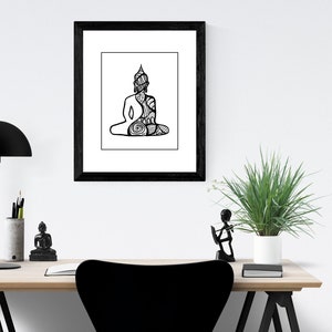 May include: Black and white line art print of a meditating Buddha in a black frame. The print is hanging on a white wall above a wooden desk with a black lamp, a small Buddha statue, and a potted plant.