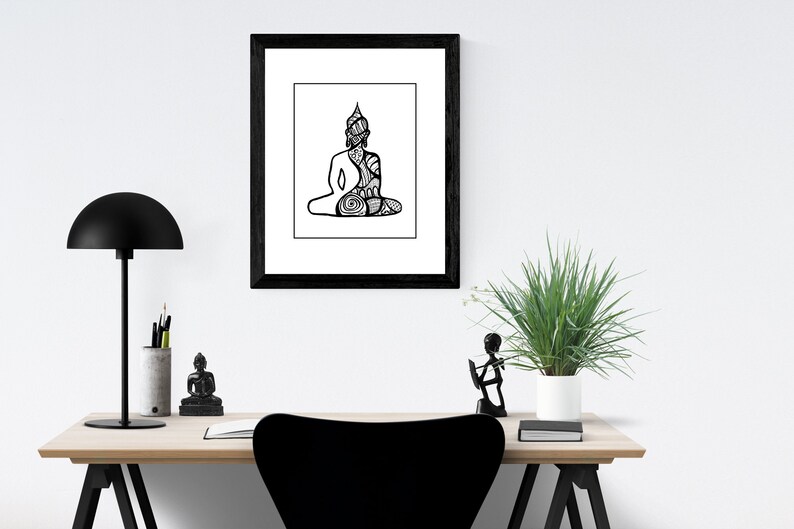 May include: Black and white line art print of a meditating Buddha in a black frame. The print is hanging on a white wall above a wooden desk with a black lamp, a small Buddha statue, and a potted plant.