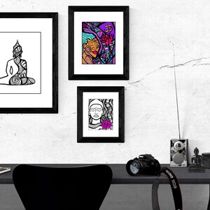 May include: Three black framed prints with white mats hanging on a white wall. The prints feature black and white line drawings of a meditating Buddha, a woman's face with a flower, and a woman's face with a flower and a symbol. A black desk with a black chair, a camera, a radio, and a small Buddha statue are in the foreground.