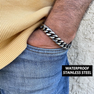 Puede incluir: Un brazalete de cadena plateado con un diseño de eslabón de bordillo. El brazalete está hecho de acero inoxidable impermeable.