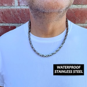 May include: A silver chain necklace made of stainless steel. The chain is a box style with a flat, rectangular link. The necklace is worn by a person with a white t-shirt. The text "WATERPROOF STAINLESS STEEL" is visible at the bottom of the image.