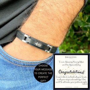 May include: A black leather bracelet with a silver cross charm. The bracelet is on a man's wrist. The image also includes a card with the text "To an Amazing Young Man on His Special Day Congratulations! Trust in the Lord with all your heart... in all your ways acknowledge Him, and He will make your paths straight. Proverbs 3:5,8"