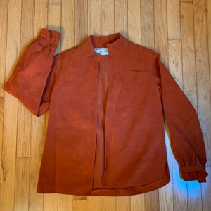 Könnte beinhalten: Rostfarbene Jacke mit Stehkragen und langen Ärmeln. Die Jacke hat eine einzelne Tasche und einen Knopfverschluss an jeder Manschette. Die offene Vorderseite zeigt das Innenfutter. Das Etikett lautet "Queen's Shop".