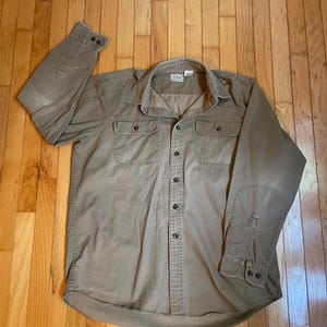 Może przedstawiać: Beżowa koszula zapinana na guziki z dwiema kieszeniami na piersi i łaty na łokciach. Koszula ma kołnierzyk i mankiety zapinane na guziki. Logo L.L.Bean jest widoczne na metce. Koszula jest wykonana z wytrzymałego materiału.