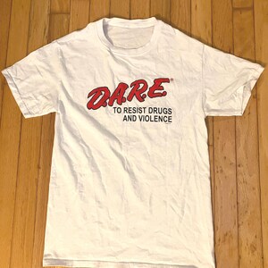 May include: A white short-sleeved t-shirt featuring the red text D.A.R.E. and the black text TO RESIST DRUGS AND VIOLENCE printed on the chest. The shirt has a crew neckline and a standard fit, displaying the iconic program logo clearly.