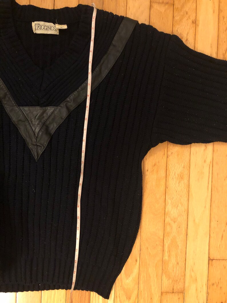 May include: A navy blue ribbed knit sweater with a V-neckline and a black leather V-shaped chest accent. A white measuring tape is placed vertically over the sweater, showing a length of approximately 28 inches from the shoulder to the hem.