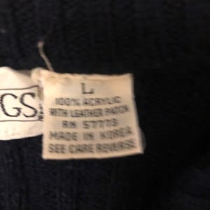 May include: A dark navy blue knit garment featuring a white fabric tag with the text L, 100% ACRYLIC WITH LEATHER PATCH, RN 57779, MADE IN KOREA, SEE CARE REVERSE, positioned next to a partial tag showing the letters GS.