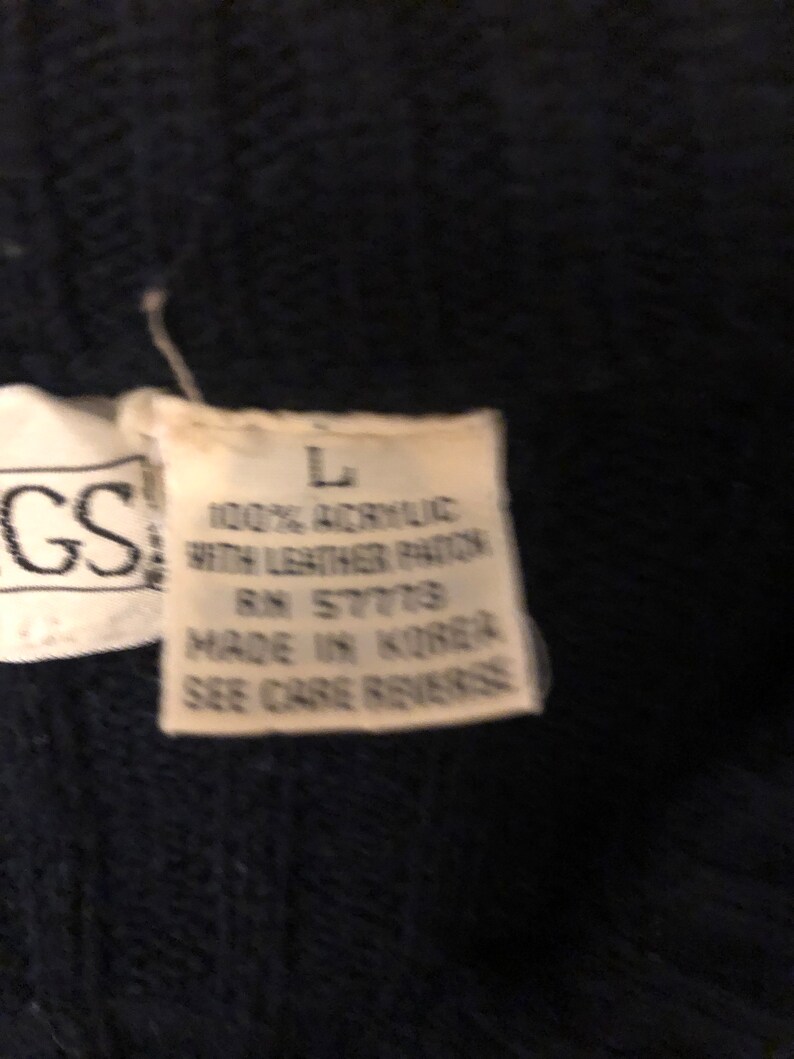 May include: A dark navy blue knit garment featuring a white fabric tag with the text L, 100% ACRYLIC WITH LEATHER PATCH, RN 57779, MADE IN KOREA, SEE CARE REVERSE, positioned next to a partial tag showing the letters GS.