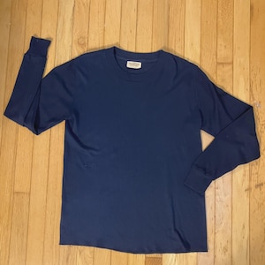 May include: A solid navy blue long-sleeve shirt featuring a crew neckline and ribbed cuffs. The garment has a straight hemline and includes a small white rectangular brand label sewn into the interior of the collar with black text.