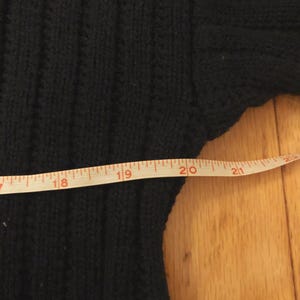 May include: A black ribbed knit sweater featuring a vertical texture pattern. A white measuring tape is positioned across the garment, showing a width measurement extending from 18 inches to just past 21 inches on the imperial scale.