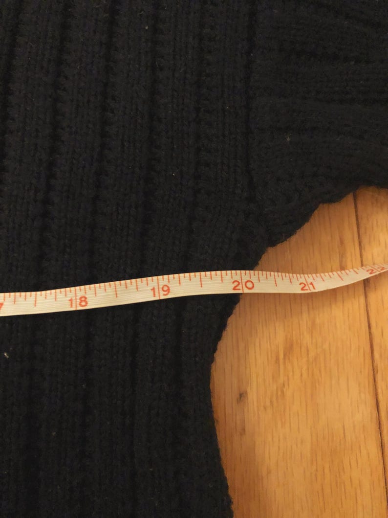 May include: A black ribbed knit sweater featuring a vertical texture pattern. A white measuring tape is positioned across the garment, showing a width measurement extending from 18 inches to just past 21 inches on the imperial scale.