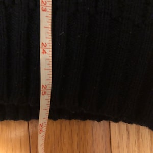 May include: A black knit sweater with a ribbed texture is positioned next to a white measuring tape. The tape shows a length measurement extending past 26 inches, indicating the vertical dimension of the garment's hemline and lower body section.