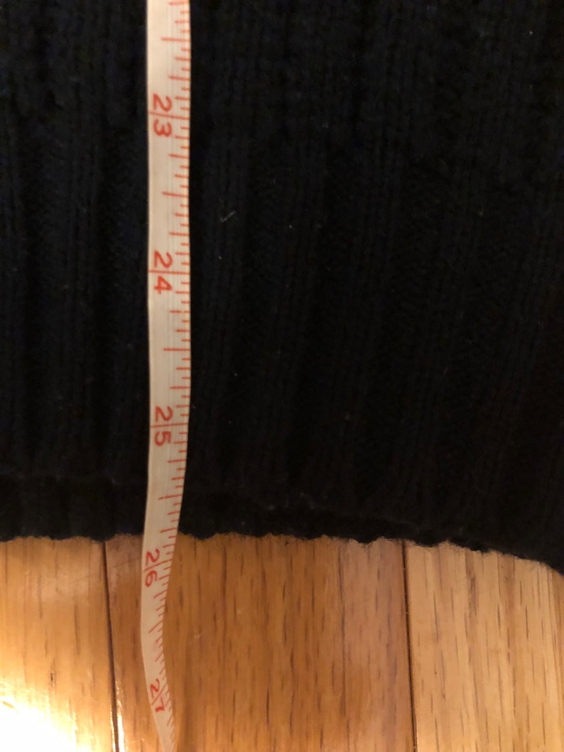 May include: A black knit sweater with a ribbed texture is positioned next to a white measuring tape. The tape shows a length measurement extending past 26 inches, indicating the vertical dimension of the garment's hemline and lower body section.