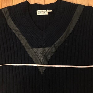 May include: A black ribbed V-neck sweater featuring black leather trim accents around the neckline. A white measuring tape is positioned across the chest, showing a width measurement of approximately 21 inches from edge to edge.