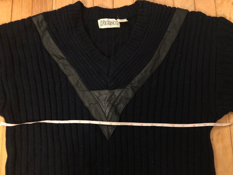 May include: A black ribbed V-neck sweater featuring black leather trim accents around the neckline. A white measuring tape is positioned across the chest, showing a width measurement of approximately 21 inches from edge to edge.