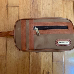 May include: A brown canvas travel toiletry bag with a brown leather trim and a zippered pocket. The bag has a brown leather handle and a Samsonite logo.