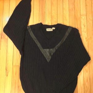 May include: A navy blue long-sleeve sweater featuring a ribbed knit texture and a V-neckline. The sweater includes a dark leather-like V-shaped accent panel across the chest and shoulders. A white brand label with the text RIGGINGS is at the neck.