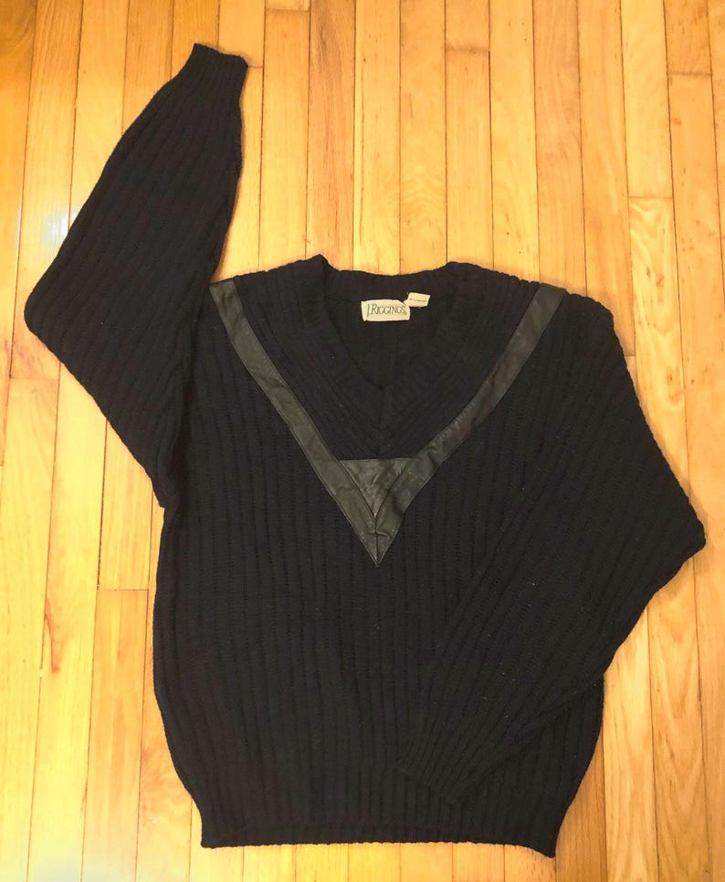 May include: A navy blue long-sleeve sweater featuring a ribbed knit texture and a V-neckline. The sweater includes a dark leather-like V-shaped accent panel across the chest and shoulders. A white brand label with the text RIGGINGS is at the neck.