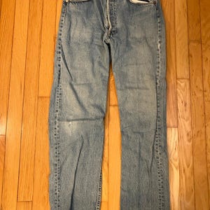 May include: A pair of light blue denim jeans featuring a classic five-pocket design, a button fly closure, and a straight-leg cut. The fabric displays a faded wash with subtle whiskering details around the hips and thighs, finished with copper-toned rivets.
