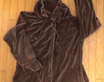 May include: A brown velvet jacket with a high, gathered collar and button-down front. The jacket has long sleeves with ribbed cuffs and a drawstring closure at the neck. The jacket is made of a soft, plush material.