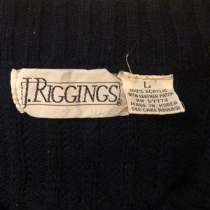 May include: A dark navy blue cable knit sweater featuring a white fabric label with the text J. RIGGINGS. A smaller adjacent label indicates size L, 100% acrylic, with leather patch, RN 57773, made in Korea, and instructions to see care reverse.