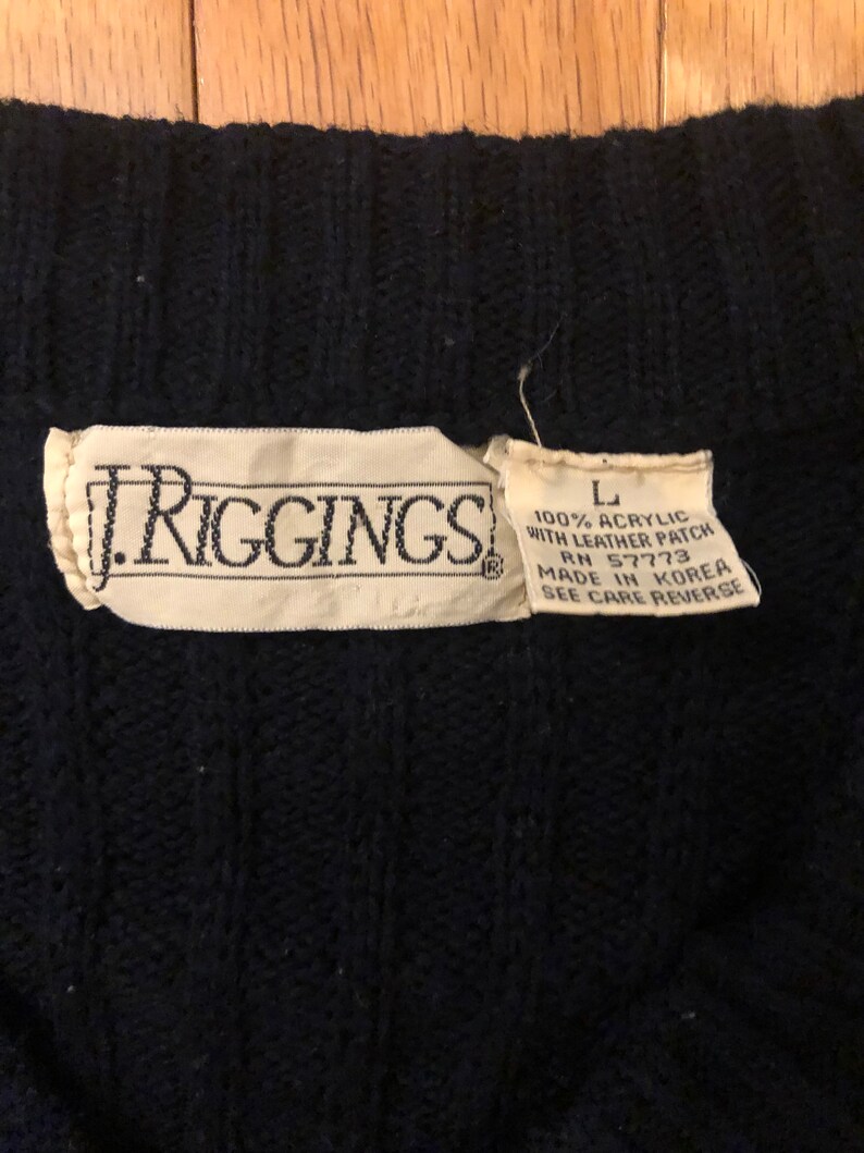 May include: A dark navy blue cable knit sweater featuring a white fabric label with the text J. RIGGINGS. A smaller adjacent label indicates size L, 100% acrylic, with leather patch, RN 57773, made in Korea, and instructions to see care reverse.