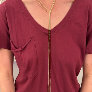 May include: A long, thin, yellow beaded lariat necklace featuring two white, round, flat coin-shaped pearls at the ends. The necklace is worn around the neck, with the two strands hanging down the center of a solid dark red v-neck t-shirt.