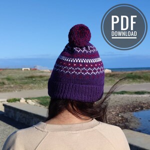 以下が含まれることがあります： 紫色のニット帽に白と赤の幾何学模様とポンポンが付いています。帽子は水辺に立っている人が被っています。