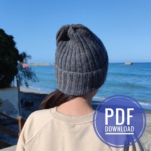 May include: A gray knitted beanie hat with a ribbed texture. The hat is worn by a person standing in front of a beach scene. The text "PDF DOWNLOAD" is visible in a purple circle.