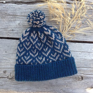 May include: A knitted blue beanie with a geometric pattern in light beige. The hat features a ribbed brim and a matching pom-pom on top. The hat is displayed on a weathered wooden surface.