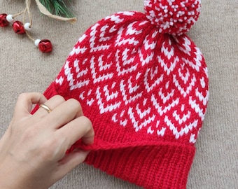 Puede incluir: Gorro de invierno de punto rojo y blanco con un pompón blanco en la parte superior. El gorro tiene un patrón geométrico.